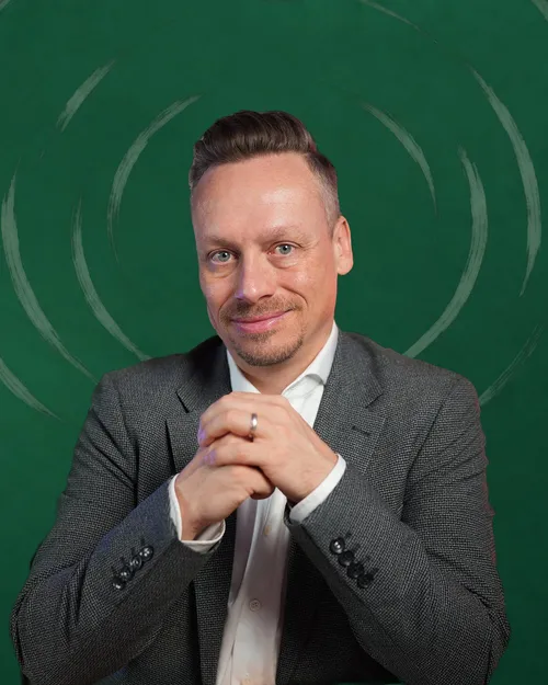 Fabian Henzler in a gray blazer, hands folded, smiling in front of forest green with drawn rings