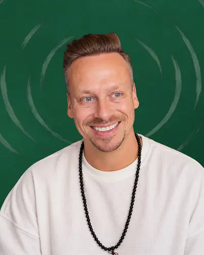 Fabian Henzler im weißen Shirt mit Mala-Kette, mit einem warmen Lächeln direkt in die Kamera, vor Waldgrün mit gezeichneten Ringen