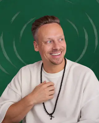Fabian Henzler in a white shirt with mala beads, laughing, his hand on his heart, in front of forest green with drawn rings