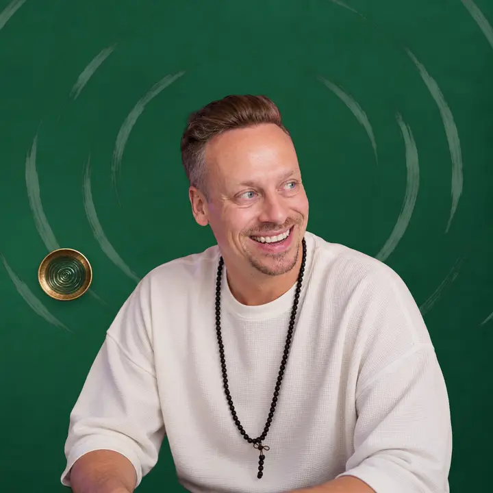 Fabian Henzler in a white shirt with mala beads, laughing and looking to the side, in front of forest green with drawn rings and a small singing bowl